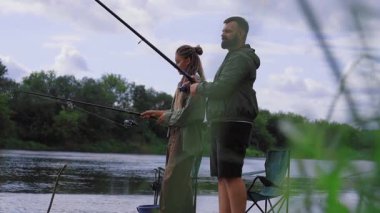 Genç çift, sakin doğa ve bulutlu bir günde yemyeşil ağaçlarla çevriliyken, ellerinde oltalarını tutarak, birlikte balık tutarak, rahatlatıcı bir günün tadını çıkarıyorlar.