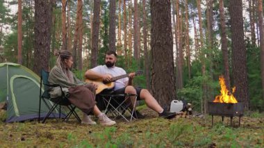 Genç romantik çift ormanda kamp yaparken, sevgilisi için akustik gitar çalan adamla birlikte, sıcak bir kamp ateşi ve çam ormanında bir çadırın yanında oturuyor.