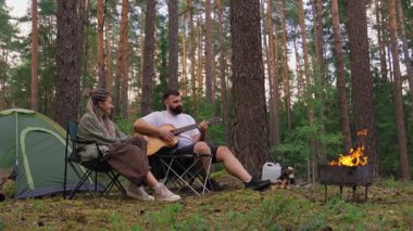 Genç romantik çift ormanda kamp yaparken, sevgilisi için akustik gitar çalan adamla birlikte, sıcak bir kamp ateşi ve çam ormanında bir çadırın yanında oturuyor.