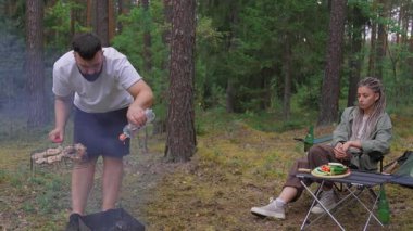 Çam ormanında kamp yapan genç bir çift: partneri kamp alanında sebze ve et pişirirken, kadın dizüstü bilgisayarla çalışıyor. Uzaktan kumandayla boş zamanı harmanlıyor.