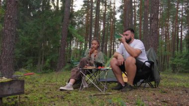 Rastalı ve sakallı beyaz bir çift ormanda ızgara et ve taze sebzelerle pikniğin tadını çıkarıyor. Seyahat, turizm ve açık hava maceralarını tanıtıyorlar.