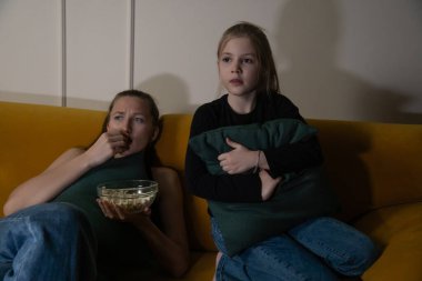 İki genç kız, loş ışıklı bir odada sarı bir kanepede oturmuş, yoğun bir şekilde korku filmi izliyorlar, biri patlamış mısır yiyor, diğeri de yastık tutuyor, korku ve gerilim ifadeleri sergiliyorlar.