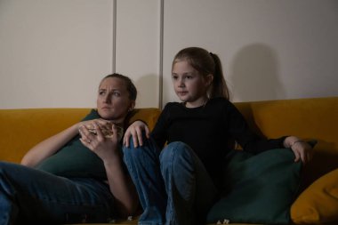 Anne ve kızı, loş oturma odasındaki sarı bir kanepede rahatlar, evde rahat bir film gecesi boyunca birlikte televizyon izlerken patlamış mısır ve yastık paylaşırlar.