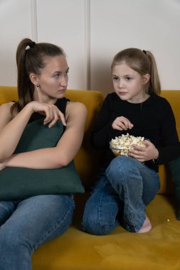 Genç bir kadın ve çocuk hardal sarısı bir kanepede birlikte oturur, ekran seyreder, bir kase patlamış mısırı paylaşır ve gevşemiş aile bağlarının tadını çıkarırlar. Dikey fotoğraf