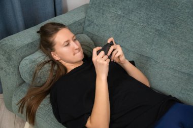 Uzun kahverengi saçlı, günlük giysiler giyen, rahat koltukta uzanan, akıllı telefonuyla mobil oyunlar oynayan, evde boş zamanlarının keyfini çıkaran genç bir kadın.