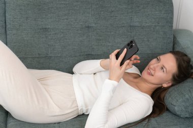 Genç bir kadın evinde rahat bir kanepeye uzanmış, akıllı telefonunu tutarak büyüleyici haberler okuyor ya da videolar seyrediyor, rahatlama ve rahatlama anının tadını çıkarıyor.