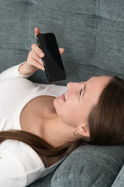 Genç bir kadın akıllı telefonunun ses asistanı özelliğini kullanarak rahat bir kanepede uzanıyor. Modern evinde rahatlık ve rahatlığın tadını çıkarıyor.