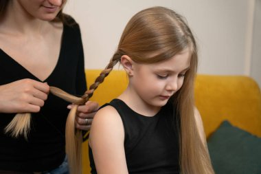 Anne, genç sarışın kızının uzun saçlarını örüp evde örüyor. Şefkatli bir ebeveyn-çocuk anı. Bakım, rutin, bağ ve günlük aile yakınlığını samimi bir ortamda gösteriyor.