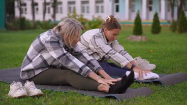 Esneklik egzersizi yapan ve yeşil bir parkta yoga minderleri üzerinde oturan iki beyaz kadın, sağlıklı bir yaşam tarzına ve dışarıda iyi bir yaşam için birlikte egzersiz yapmaya odaklanıyor.