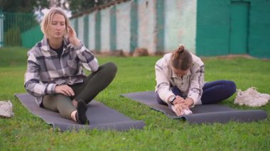 Esneklik egzersizi yapan ve yeşil bir parkta yoga minderleri üzerinde oturan iki beyaz kadın, sağlıklı bir yaşam tarzına ve dışarıda iyi bir yaşam için birlikte egzersiz yapmaya odaklanıyor.