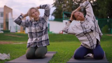 İki atletik kadın gündelik giysiler içinde eşzamanlı boyun ve omuz egzersizleri yaparken yeşil çimlerin üzerinde yoga minderleri üzerinde diz çökerek sağlıklı ve aktif bir yaşam tarzını teşvik ediyorlar.