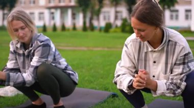 İki atletik kadın, sabah antrenmanlarından önce esneklik ve esneme üzerine odaklanarak yeşil bir parkta yoga minderleri üzerinde esneme egzersizleri ve ısınma hareketleri yapıyorlar.