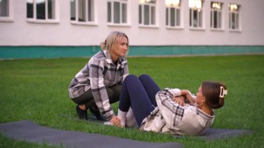 Genç atletik bir kadın yoga minderinde abdominal mekik çekerken kadın bir fitness eğitmeni yeşil çimlerde motivasyon ve rehberlik sağlar.