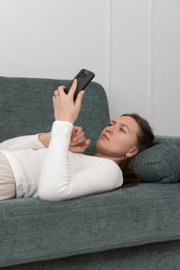 Genç kadın evdeki rahat kanepede uzanıyor, akıllı telefon kullanıyor, internette geziniyor, sosyal medyada sohbet ediyor, video ve fotoğraf izliyor.