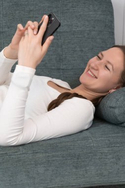 Genç kadın evdeki rahat kanepede uzanıyor, akıllı telefon kullanıyor, internette geziniyor, sosyal medyada sohbet ediyor, video ve fotoğraf izliyor, gülümsüyor ve boş zamanlarının tadını çıkarıyor. Dikey fotoğraf