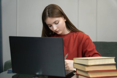 Genç kız dizüstü bilgisayar kullanırken, not alırken ve evde çalışırken, çevrimiçi öğrenim, uzaktan çalışma veya eğitim kavramını gösterirken, konsantre oluyor.