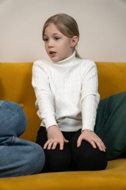 Sarı saçlı ve beyaz balıkçı yaka kazaklı genç bir kız dikkatlice dinliyor ve yeşil yastıklı hardal sarısı bir kanepede otururken konuşuyor, tartışmaya odaklanıyor. Dikey fotoğraf
