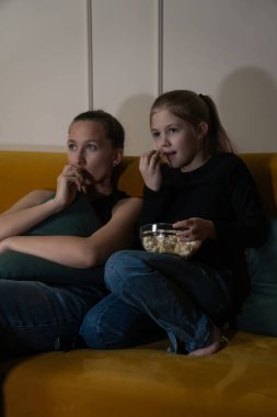 İki kız, sarı bir kanepede oturup ciddi ifadelerle televizyon seyrediyor. Biri yastığa sarılıyor, diğeri de loş bir odada elinde patlamış mısır tutuyor. Dikey fotoğraf