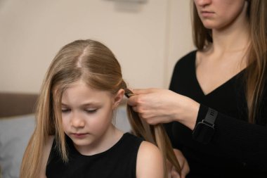 Anne, genç kızının uzun sarı saçlarını dikkatlice örüyor, düzenli bir at kuyruğu yapıyor, ebeveynler ve çocuklar arasındaki ilgiyi, sevgiyi ve aile bağlarını simgeliyor.