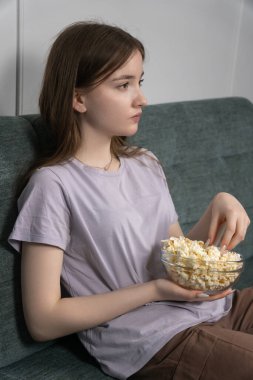 Genç kız boş zamanlarında film izleyip patlamış mısır yerken evinde tipik bir rahatlama ve eğlence sahnesini temsil ederek oturma odasında rahat bir şekilde oturuyor.