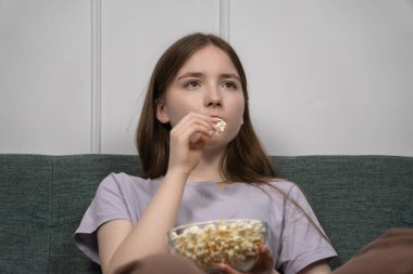 Genç kız rahat bir kanepede oturmuş, bir kase patlamış mısırla film gecesinin tadını çıkarıyor, evde boş vakit ve eğlence anı yaşıyor.