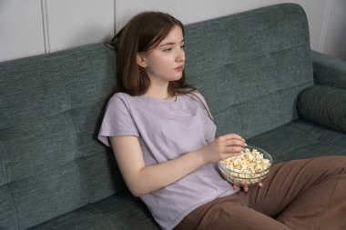 Sıkıcı genç kız sıcak bir kanepede uzanmış elinde patlamış mısırla oturma odasında sıkıcı bir TV şovu izlerken can sıkıntısının ve ilgisizliğin ağırlığını hissediyor.