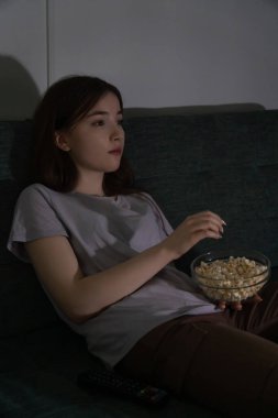 Rahat bir genç kız evinde rahat bir akşamın tadını çıkarıyor, kanepede oturuyor, televizyon izliyor ve yanında uzaktan kumandayla cam bir kaseden patlamış mısır yiyor.