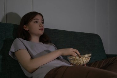 Genç kız geceleri oturma odasında rahat bir kanepede uzanıyor cam kaseden patlamış mısır yiyor ve televizyonda film izliyor.