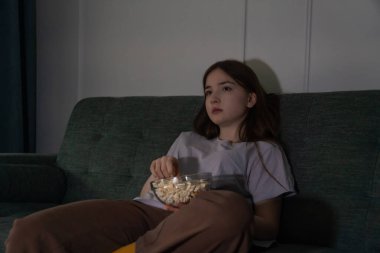 Düşünceli genç kız televizyon seyredip patlamış mısırın tadını çıkarırken loş ışıklı bir oturma odasında dinleniyor ve evdeki sessiz bir gecede huzurlu bir atmosfer yaratıyor.