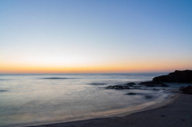 Deniz sakin ve gökyüzü turuncu. Güneş batıyor ve su gökyüzünün renklerini yansıtıyor.