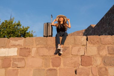 Güneşli bir günde seyahat valiziyle oturan genç bir bayan turist. Seyahat, yaz tatili ve tatil konsepti.