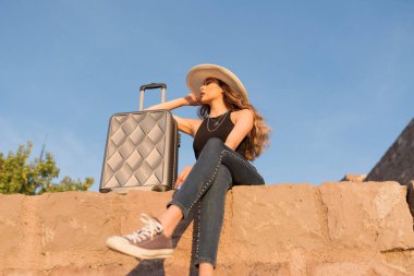 Güneşli bir günde seyahat valiziyle oturan genç bir bayan turist. Seyahat, yaz tatili ve tatil konsepti.