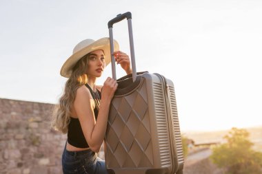 Güneşli bir günde seyahat valiziyle oturan genç bir bayan turist. Seyahat, yaz tatili ve tatil konsepti.