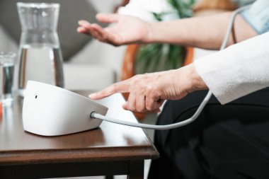 Evde dijital monitör kullanarak kan basıncını ölçen yaşlı bir kadın. Sağlık, hipertansiyon kontrolü, kendini izleme ve sağlıklı yaşlanma kavramı.