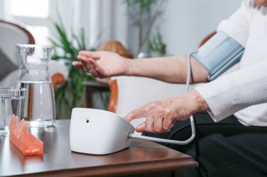 Evde dijital monitör kullanarak kan basıncını ölçen yaşlı bir kadın. Sağlık, hipertansiyon kontrolü, kendini izleme ve sağlıklı yaşlanma kavramı.