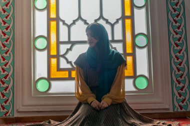 Genç Müslüman bir kadın caminin içinde renkli bir vitrinin yanında dua ediyor. Huzurlu ruhani bir an. İslam ibadeti, inanç ve bağlılık. Sakin, kutsal bir iç mekanda..