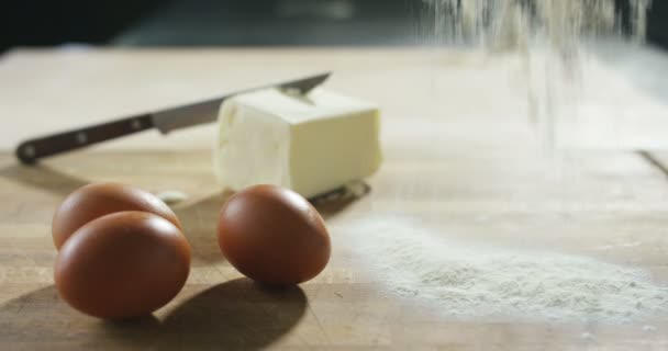 Мука падает в крайнем замедленном темпе на деревянной поверхности на кухне с фоновой палочкой масла и рядом с тремя свежими куриными яйцами — стоковое видео