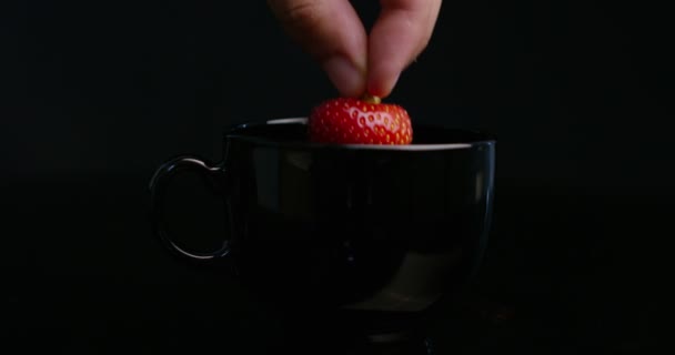 Movimento super lento ainda vida de mão jovem gotejamento de um morango em um chocolate quente derramado em uma caneca de porcelana preta isolada em um fundo preto em 4k (macro close-up ) — Vídeo de Stock