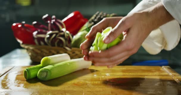 Mãos cuidadas de um jovem chef cortar suavemente em movimento lento extremo um pequeno grupo de vazamentos em uma placa de corte de madeira com fundo vegetal — Vídeo de Stock