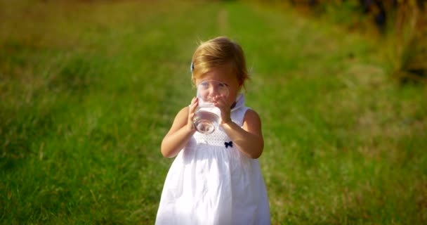 Linda menina cerca de dois anos imerso na natureza, vestido limpo beber um copo de água doce cristalina — Vídeo de Stock