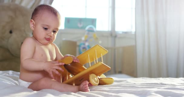 Een schattige kleine baby kijkt rond en spelen met een houten speelgoed vliegtuig — Stockvideo