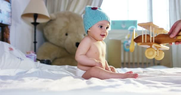 Un pequeño bebé lindo está mirando a la cámara y lleva un sombrero azul claro, su padre juega con un avión de juguete de madera — Vídeos de Stock