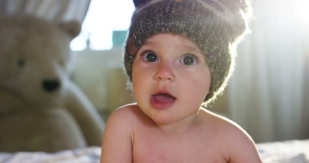 かわいい赤ちゃんがカメラに探しているし、茶色の帽子を着ている、赤ちゃんの周りに見える腕と遊ぶ — ストック動画