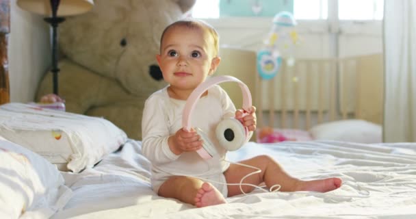 Um bebezinho bonito está olhando para a câmera e está usando um fone de ouvido rosa O bebê olha em volta e depois sorri e ri . — Vídeo de Stock