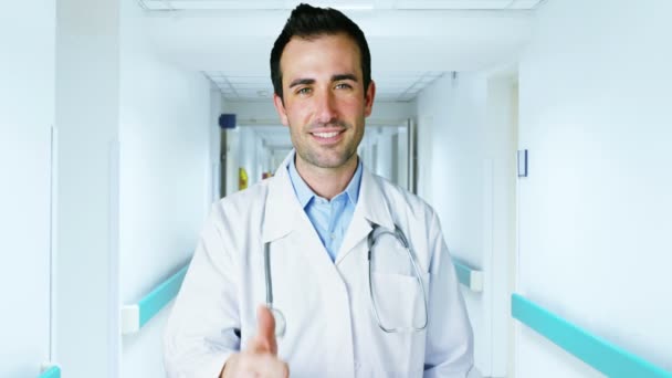 Γοητευτικό νεαρό γιατρό που κοιτάζει τη φωτογραφική μηχανή σε ένα κομψό κλινική διάδρομο χαμογελαστός και δίνοντας μια έκφραση ασφαλείας στη δουλειά του — Αρχείο Βίντεο