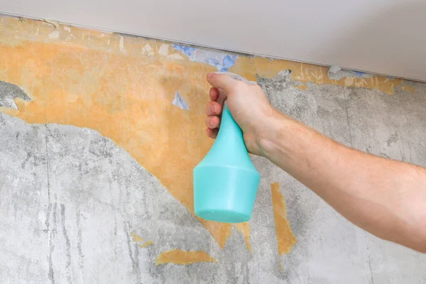 Um homem remove papel de parede velho com uma espátula e spray garrafa com água. — Fotografia de Stock