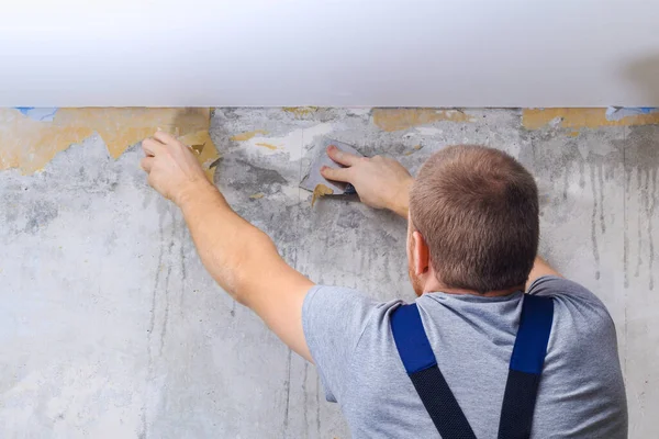 Um homem remove papel de parede velho com uma espátula e spray garrafa com água. — Fotografia de Stock