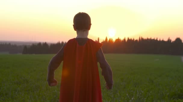 Rapaz com uma capa de super-homem num campo durante o pôr-do-sol — Vídeo de Stock