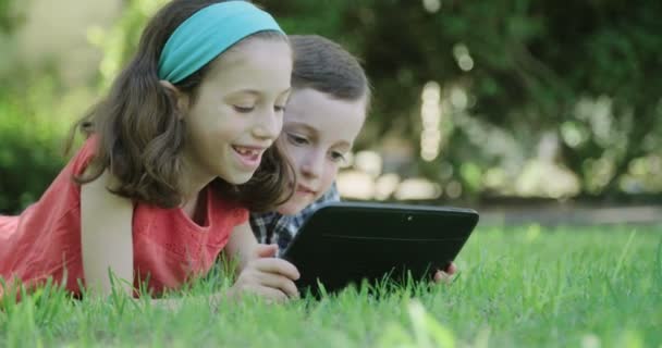 Un niño y una niña tendidos en el césped jugando con una tableta — Vídeo de stock