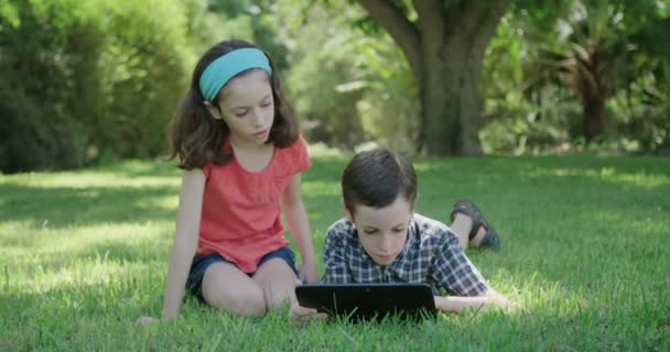 Мальчик и девочка, лежащие на газоне и играющие с планшетным компьютером — стоковое видео